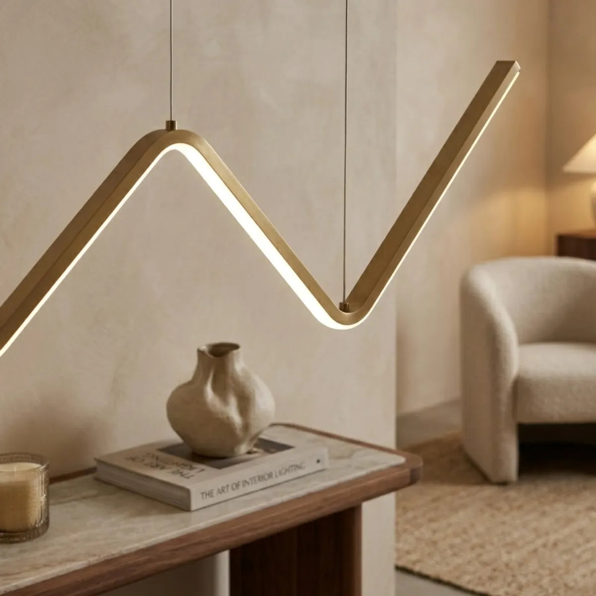 Luminária pendente dourada em formato de zigue-zague, vista em ângulo aproximado. Ela ilumina um aparador estreito com tampo de pedra, onde repousam um vaso de design orgânico e um livro de decoração. Ao fundo, uma poltrona bege desfocada.