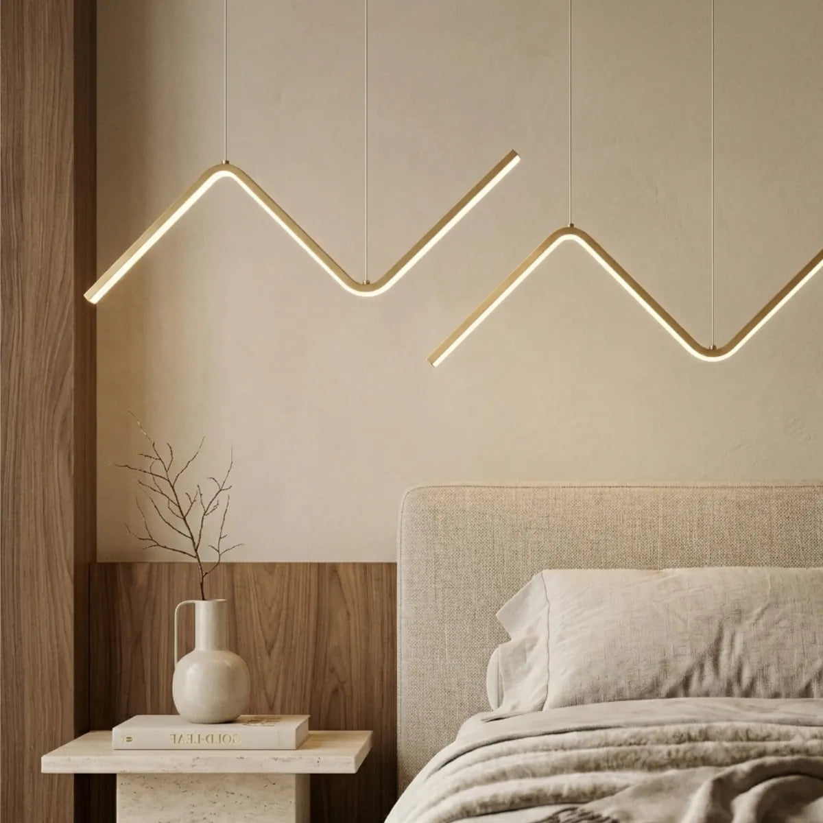Duas luminárias pendentes douradas em formato de zigue-zague iluminando um quarto de casal. Elas estão suspensas sobre uma cama com cabeceira e roupas de cama em tons de bege claro. À esquerda, uma mesa de cabeceira de pedra com um vaso branco decorativo em frente a um painel de madeira.