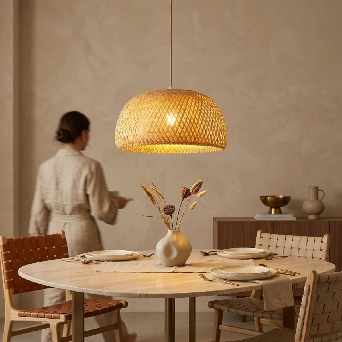 Luminária Pendente Bambu Trama suspensa sobre uma mesa de jantar redonda de madeira com lugares postos, em um ambiente rústico e elegante com uma pessoa ao fundo.