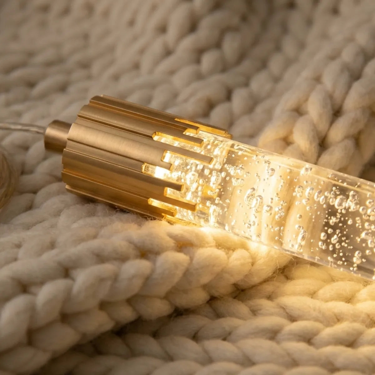 Close-up da luminária Feixe sobre manta de tricô, destacando a textura do cristal com bolhas e o design do metal dourado.