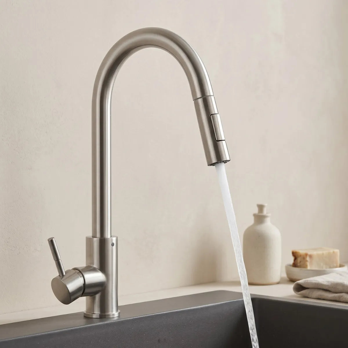 Torneira para cozinha monocomando inox com bica extensível em uso, jato d'água direcionado sobre cuba preta, fundo neutro com saboneteira e sabão artesanal.